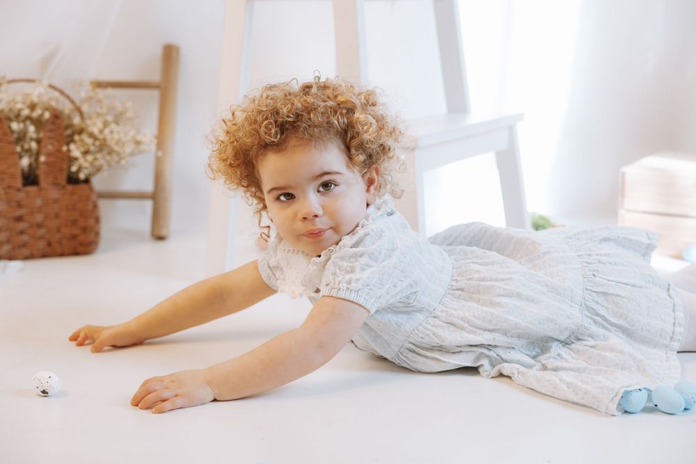 A young child in a light blue dress with curly hair lies on a white floor in a bright, airy room with decorative details.