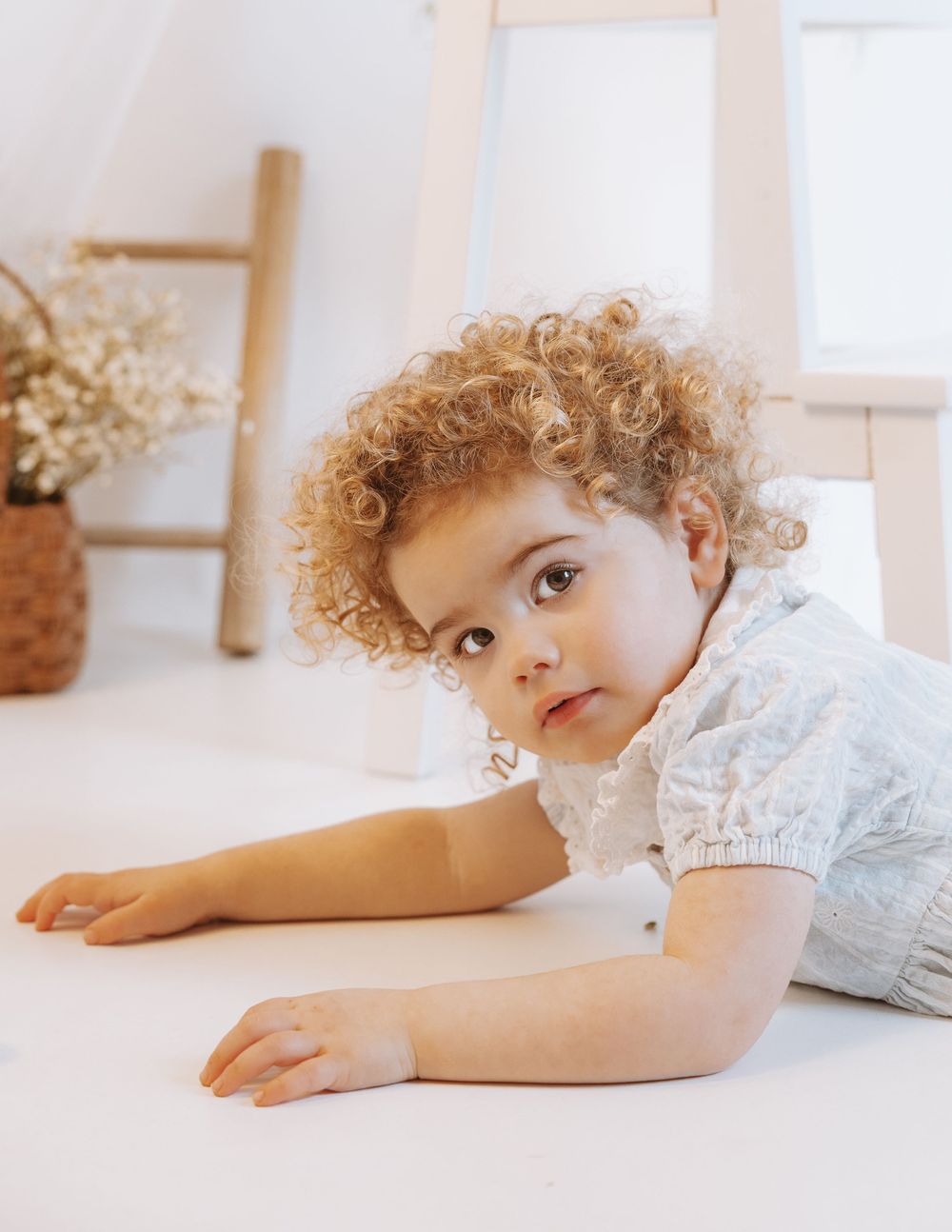 A young child in a white lacy outfit lies on a light surface near a decorative basket with dried flowers.