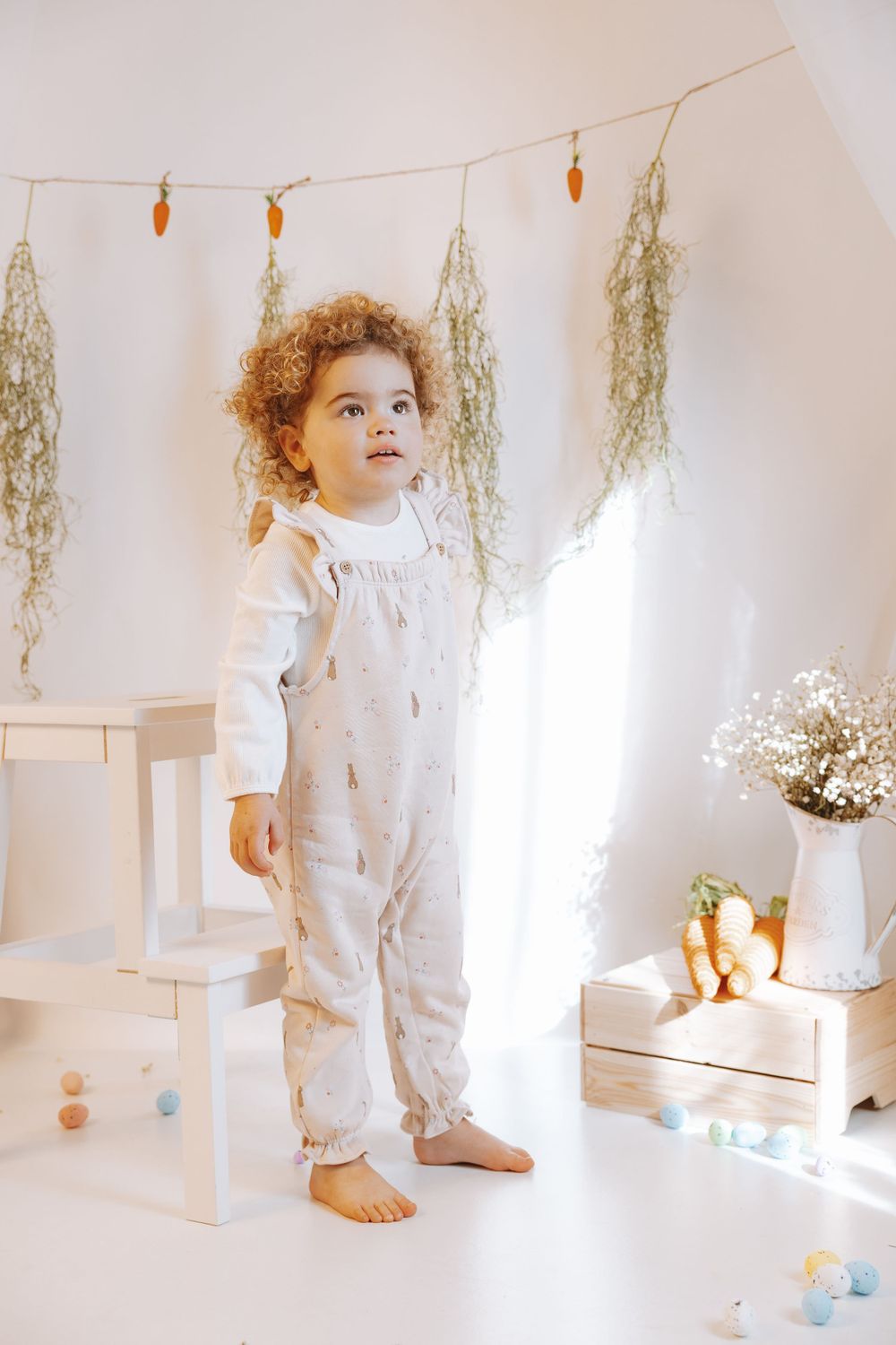 Small child wearing white overalls and shirt stands barefoot in bright minimalist room with dried flowers and decorative elements.