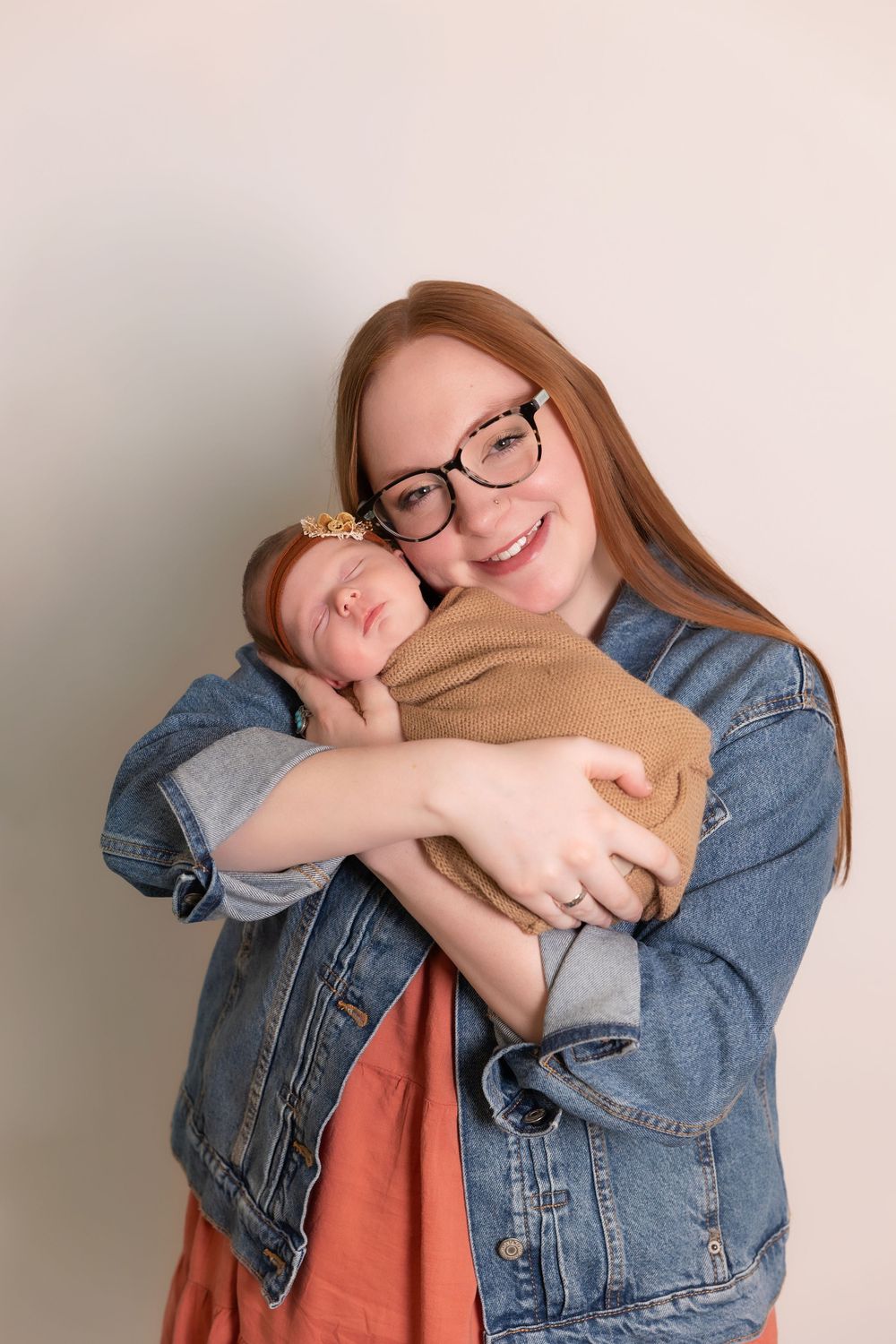 A woman wearing glasses and denim jacket tenderly embraces a sleeping baby in beige clothing against a neutral wall.