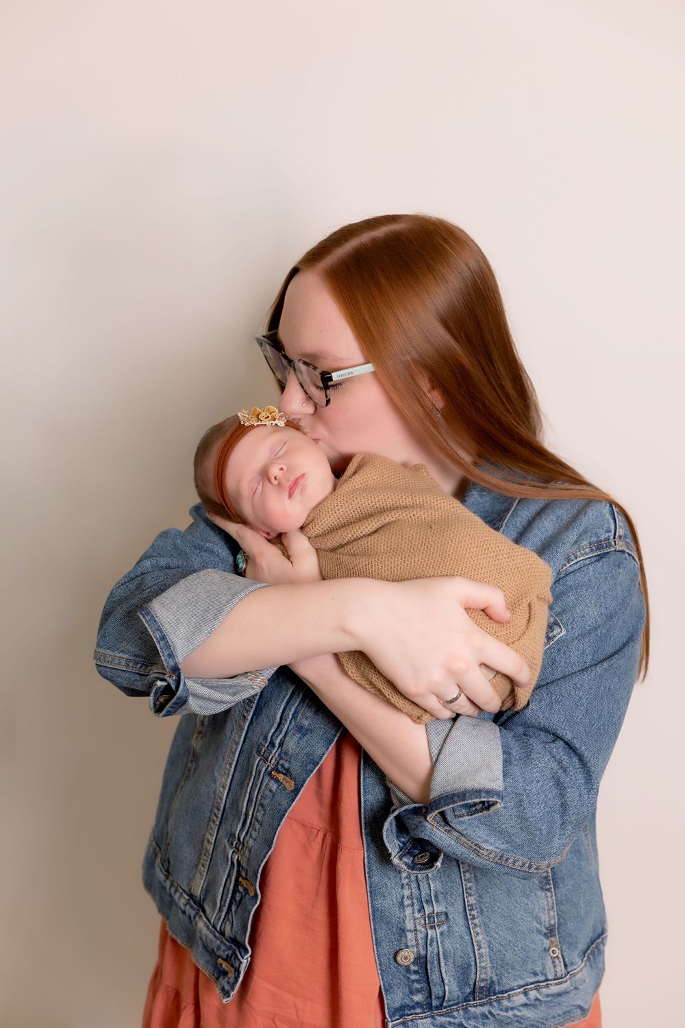A tender moment captured as a woman in denim jacket holds a sleeping infant wrapped in brown against a light background.