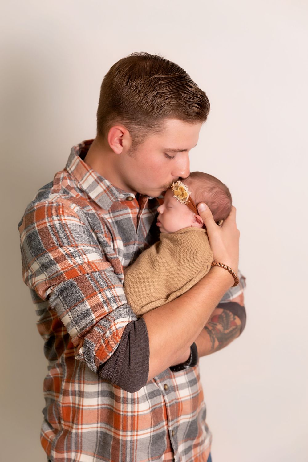 A tender moment as someone in a plaid flannel shirt holds and kisses a small baby wearing a brown sweater.