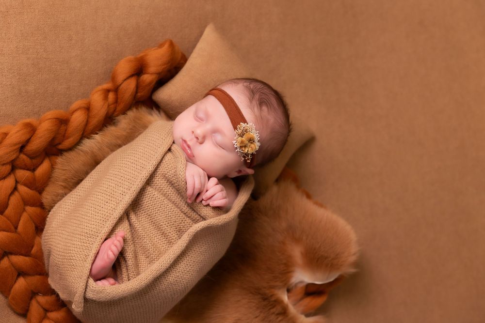 Newborn baby sleeps peacefully on brown backdrop wrapped in cozy knitted blanket with braided edge detail.