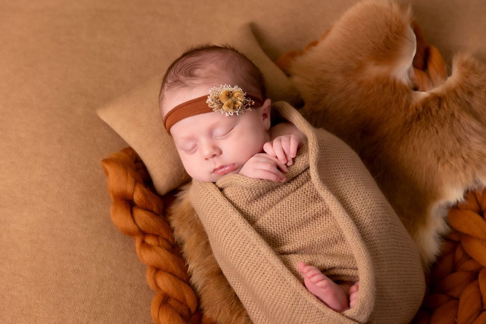 A sleeping newborn swaddled in brown cloth with a delicate floral headband rests on an autumn-toned backdrop.