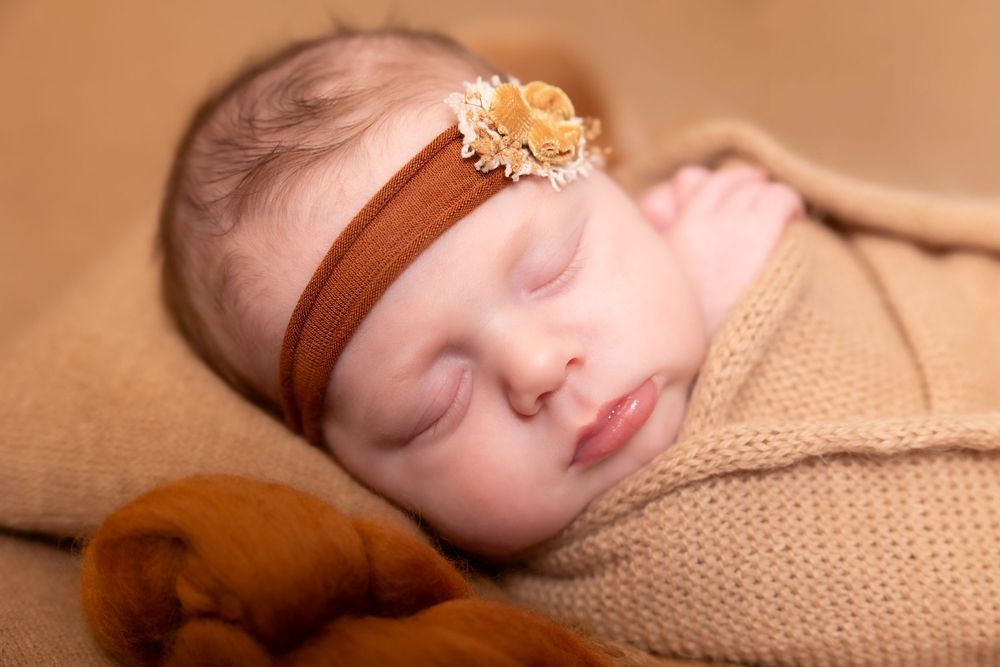 Sleeping newborn in tan wrap wearing brown headband with flower on brown backdrop.
