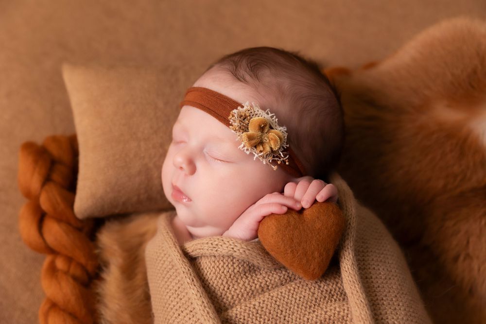 Sleeping infant wrapped in brown fabric with delicate floral headband resting on brown textured backdrop.