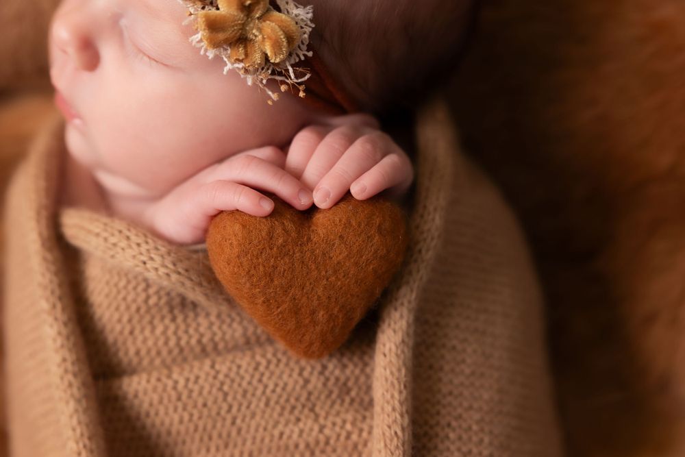 Tiny hands hold a brown heart-shaped decoration against a soft knit blanket in warm muted tones.