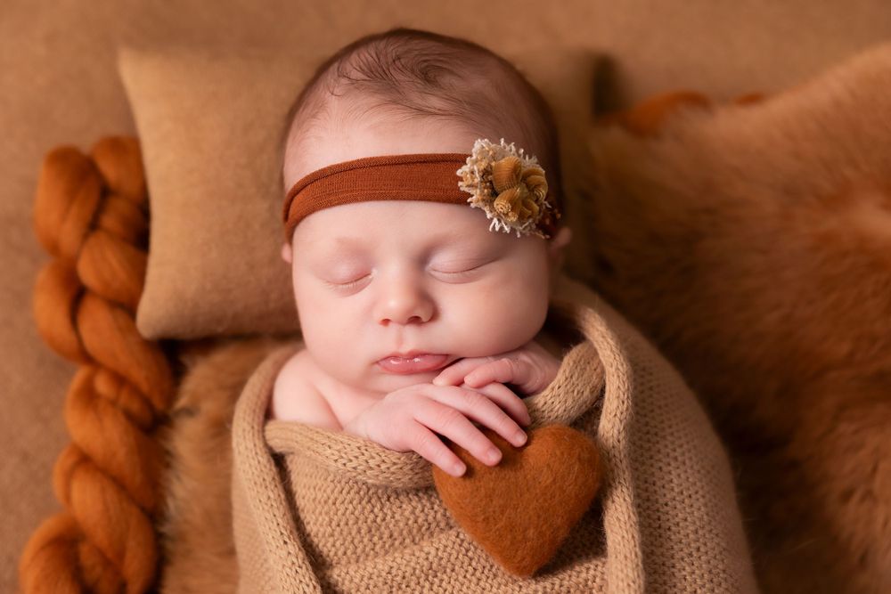 Sleeping newborn baby wrapped in brown knitted blanket with rustic orange headband resting on textured backdrop.