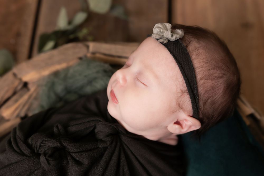 Sleeping newborn wearing black headband rests peacefully on dark fabric with rustic wooden background.