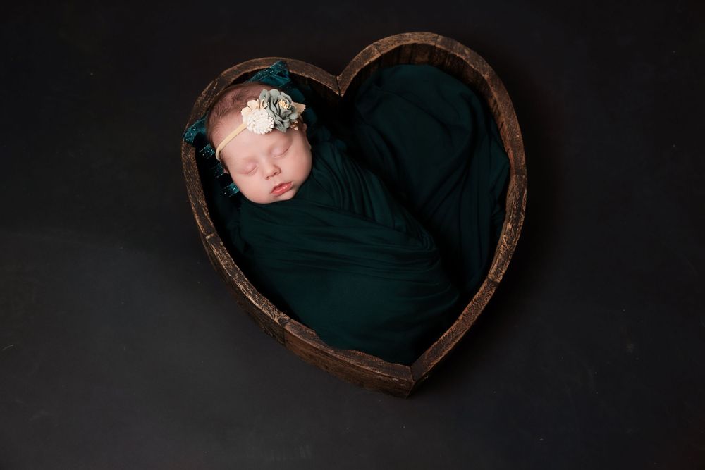 Sleeping newborn wrapped in dark teal fabric lies in heart-shaped wooden basket against black background.