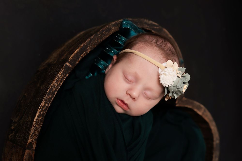 Peaceful newborn sleeps in black wrap with teal and white floral headband against dark backdrop.