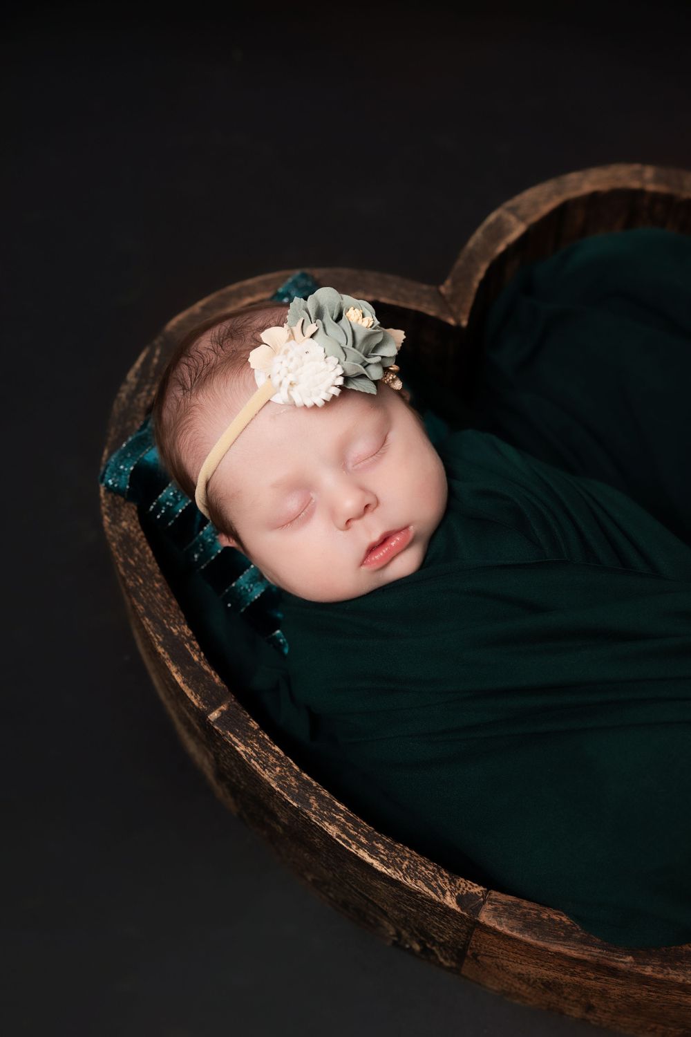 Sleeping newborn wrapped in dark teal fabric lying in rustic wooden bowl with delicate floral headband.