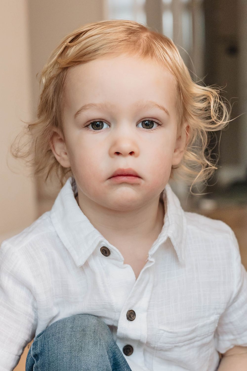 Adorable blonde toddler boy with curly hair in white linen shirt looking at camera.