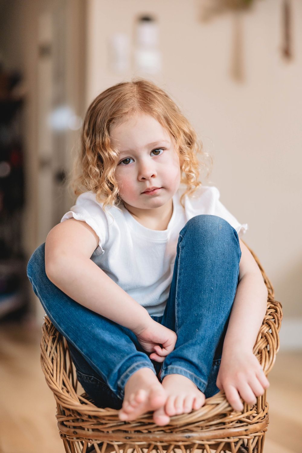 Young girl with curly red hair sits cross-legged in a wicker chair, wearing white t-shirt and jeans.