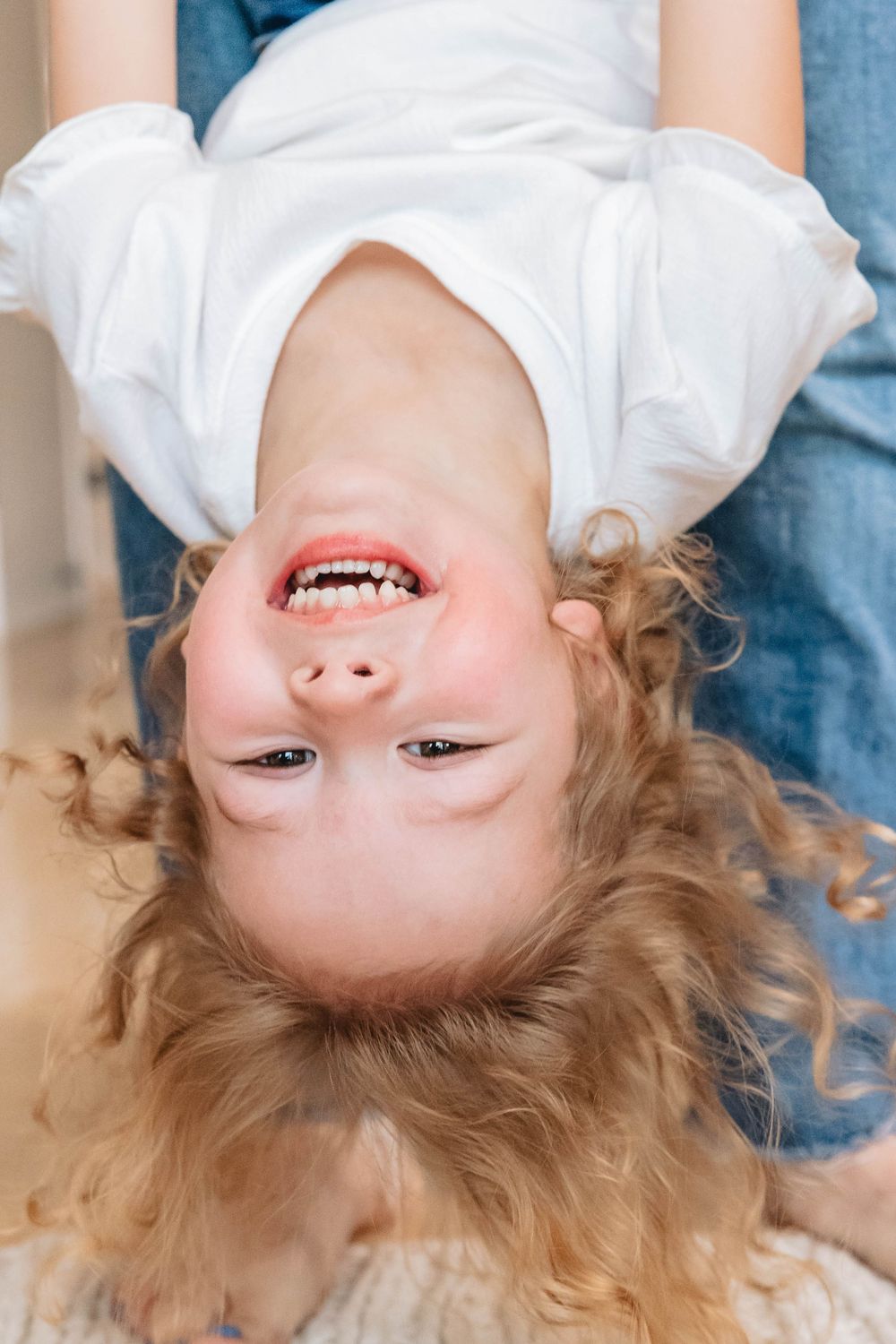 Happy young girl with curly blonde hair laughing upside down, wearing a white t-shirt and jeans.