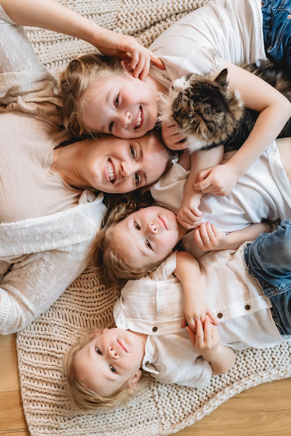 Mother and three young children lying on a knit blanket smiling with a tabby cat overhead view.