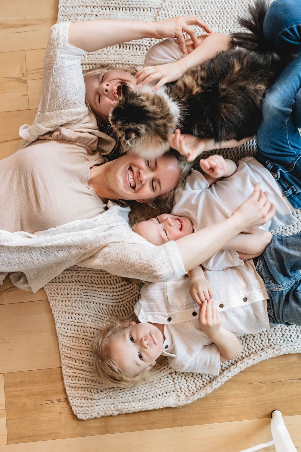 Overhead view of joyful family with mother, three kids, and tabby cat lying on knit blanket on floor.