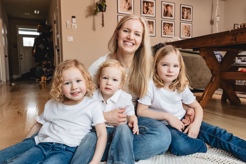 Smiling blonde mother sitting on floor with three young children in matching white shirts and jeans.