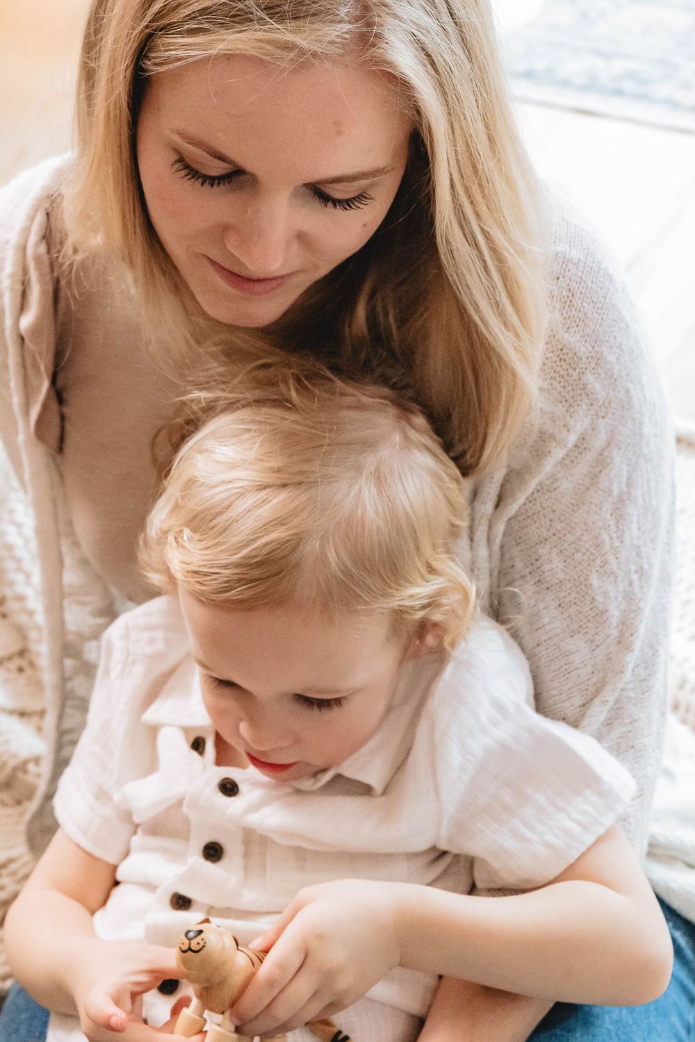 Blonde mother in cream sweater tenderly holding young toddler in white shirt on her lap indoors.