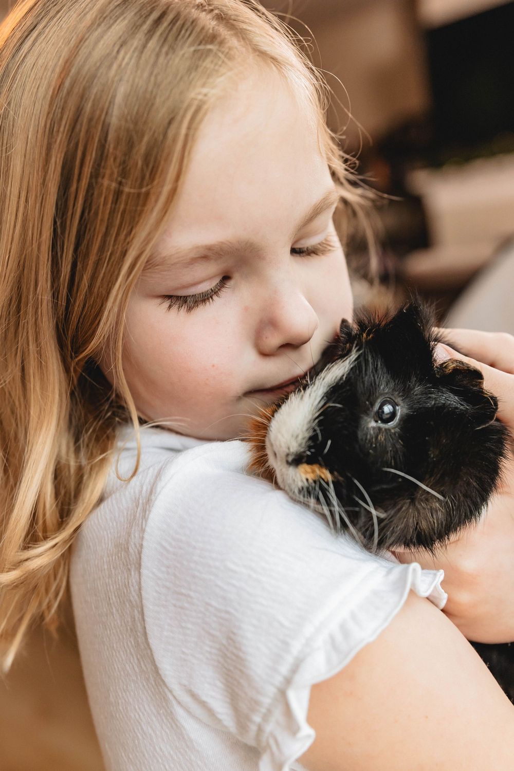 Young blonde girl with closed eyes gently kissing a black and white guinea pig she holds lovingly.