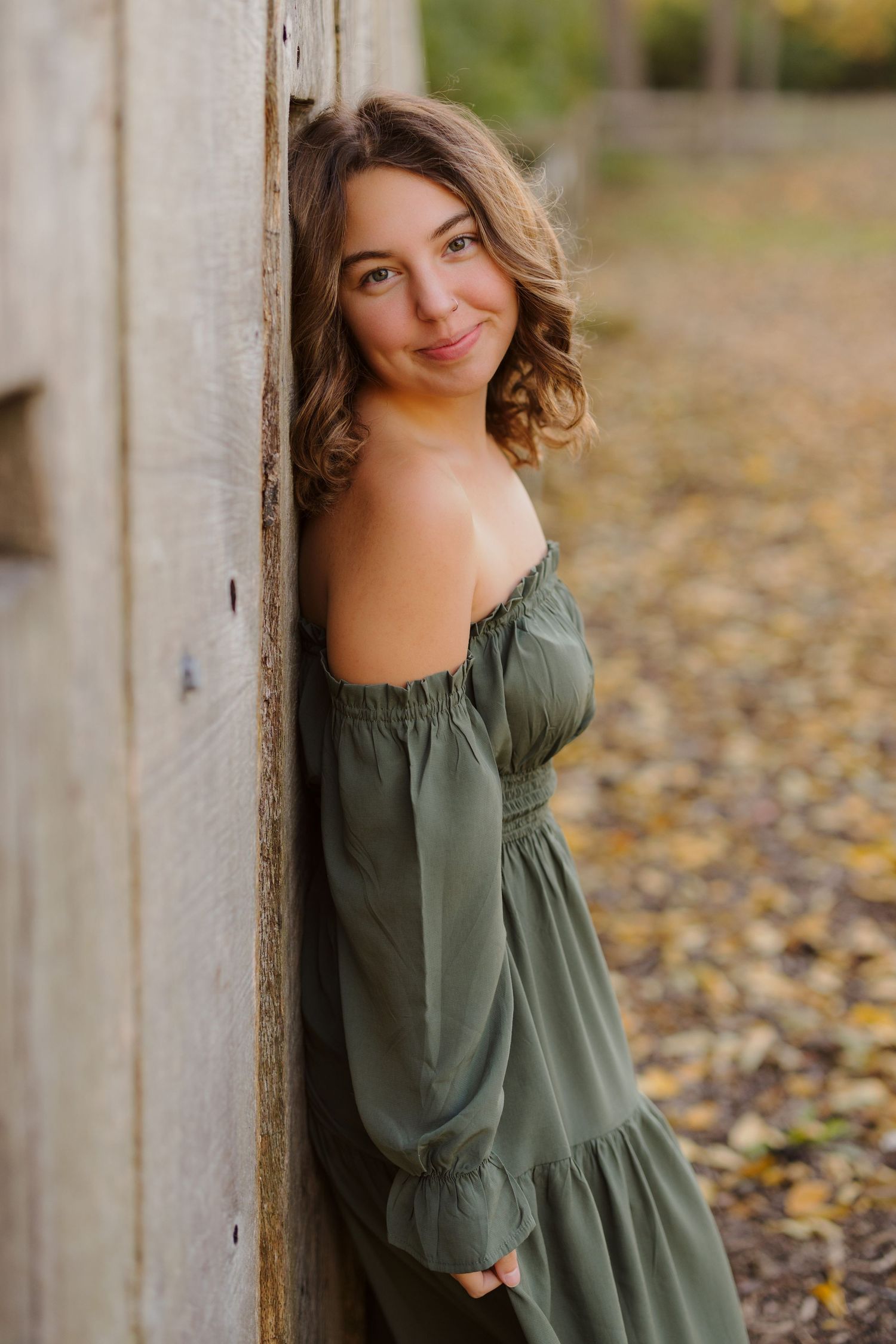 An autumnal outdoor portrait of a person in an olive green dress leaning against a rustic wall with fall leaves on the ground.