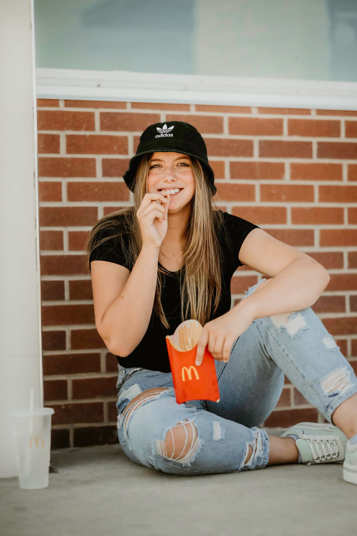Person in ripped jeans and Adidas hat sits casually against brick wall while enjoying McDonald's french fries.