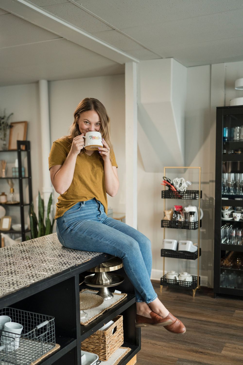 Someone in casual attire sits on a counter drinking from a mug in a modern home interior with shelving.