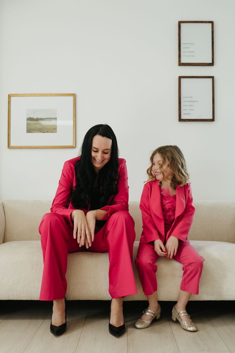 Woman and girl in matching hot pink suits sitting on a beige sofa, smiling and laughing together.