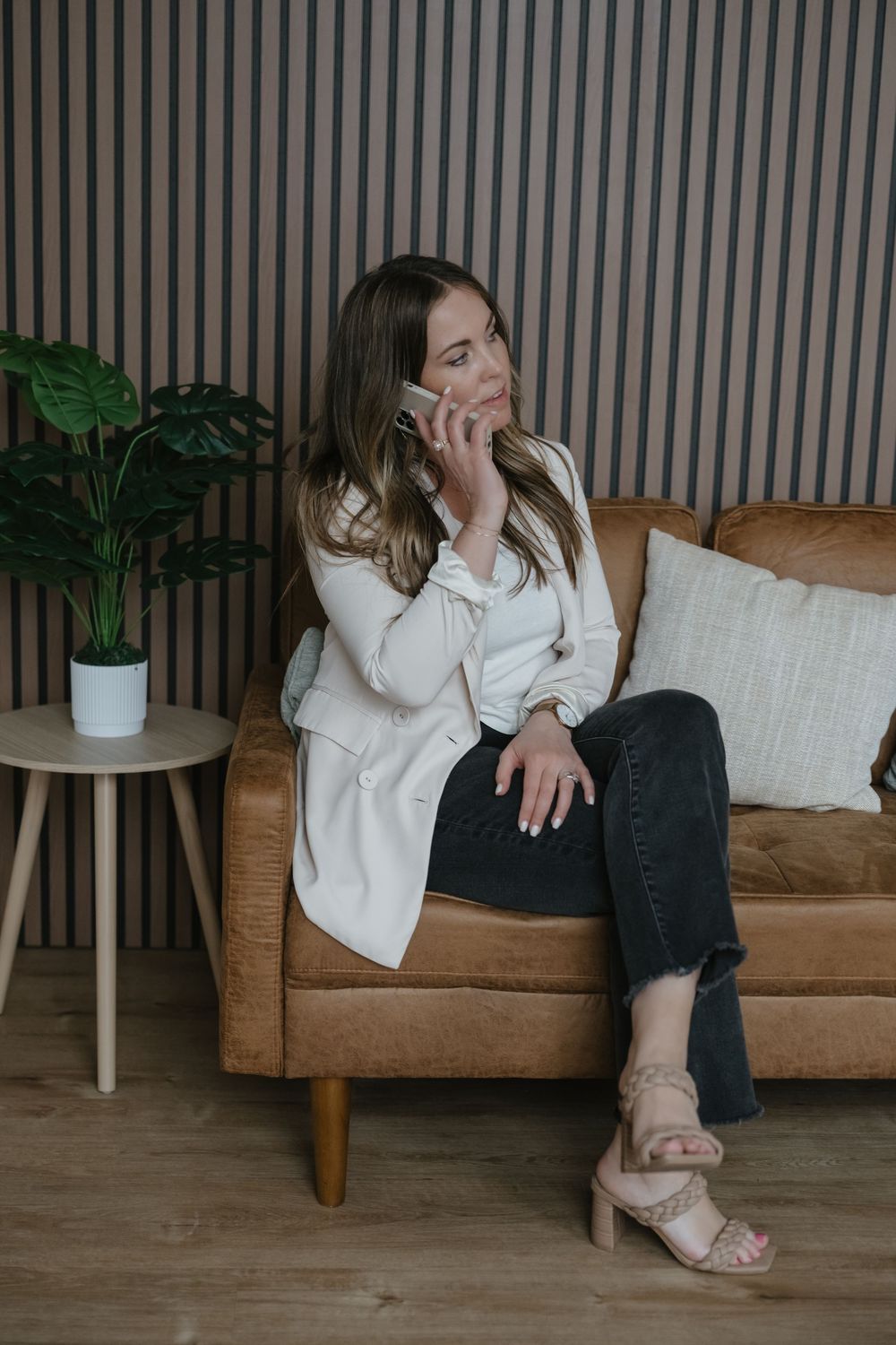 Woman in white blazer and black jeans sitting on brown leather sofa talking on phone.