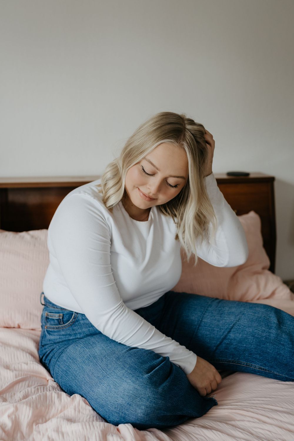 Someone in casual attire with blonde hair sits relaxed on a bed with pink bedding and denim jeans.