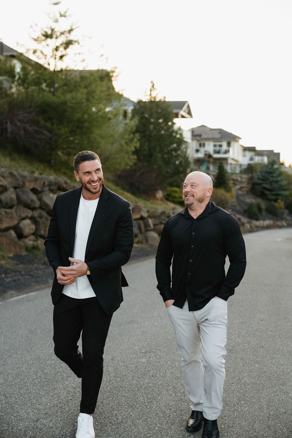 Two smiling men in black outfits walk together on a suburban road surrounded by trees and houses.