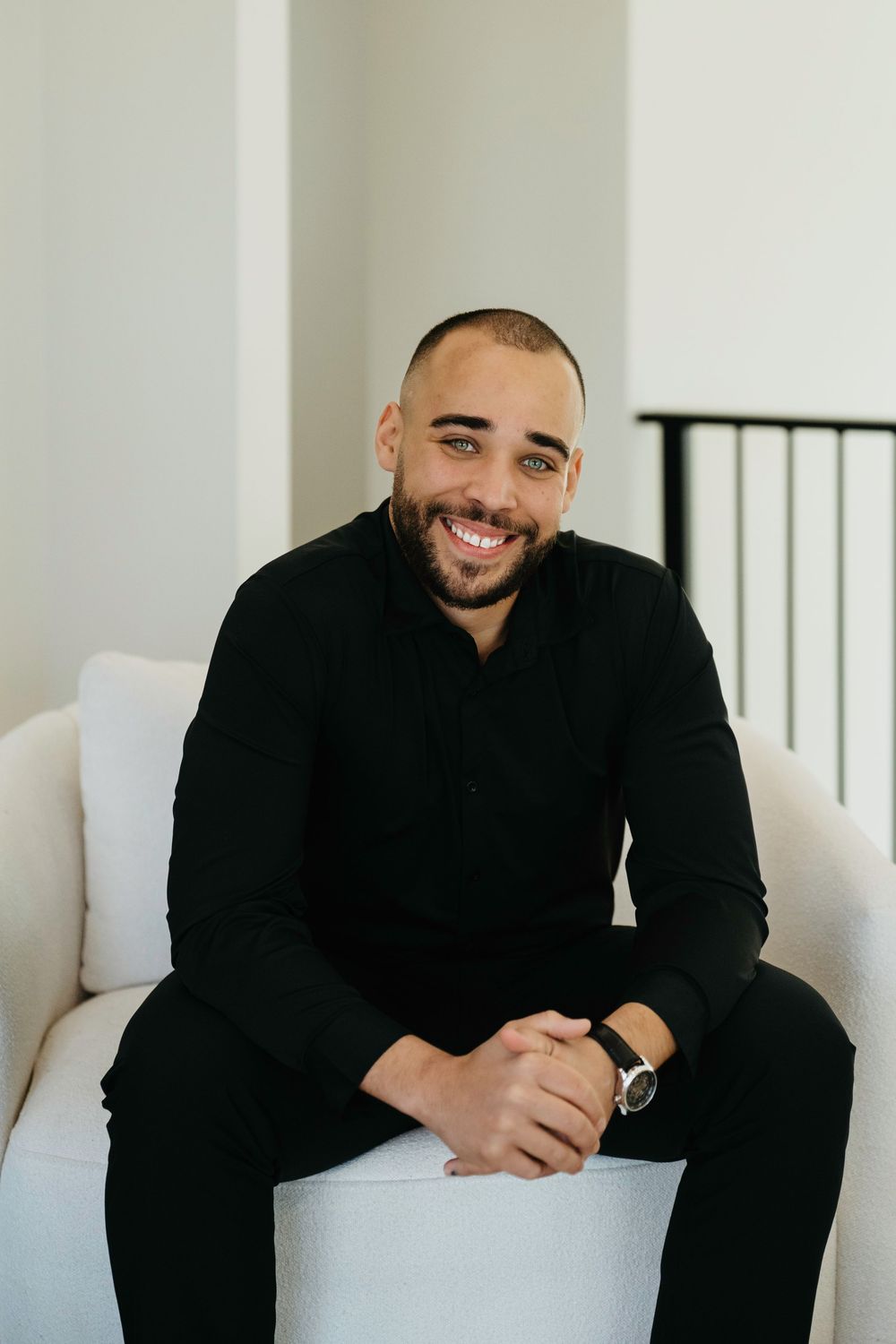 Smiling man in black outfit sitting relaxed on white chair, wearing a watch, in a bright modern interior.
