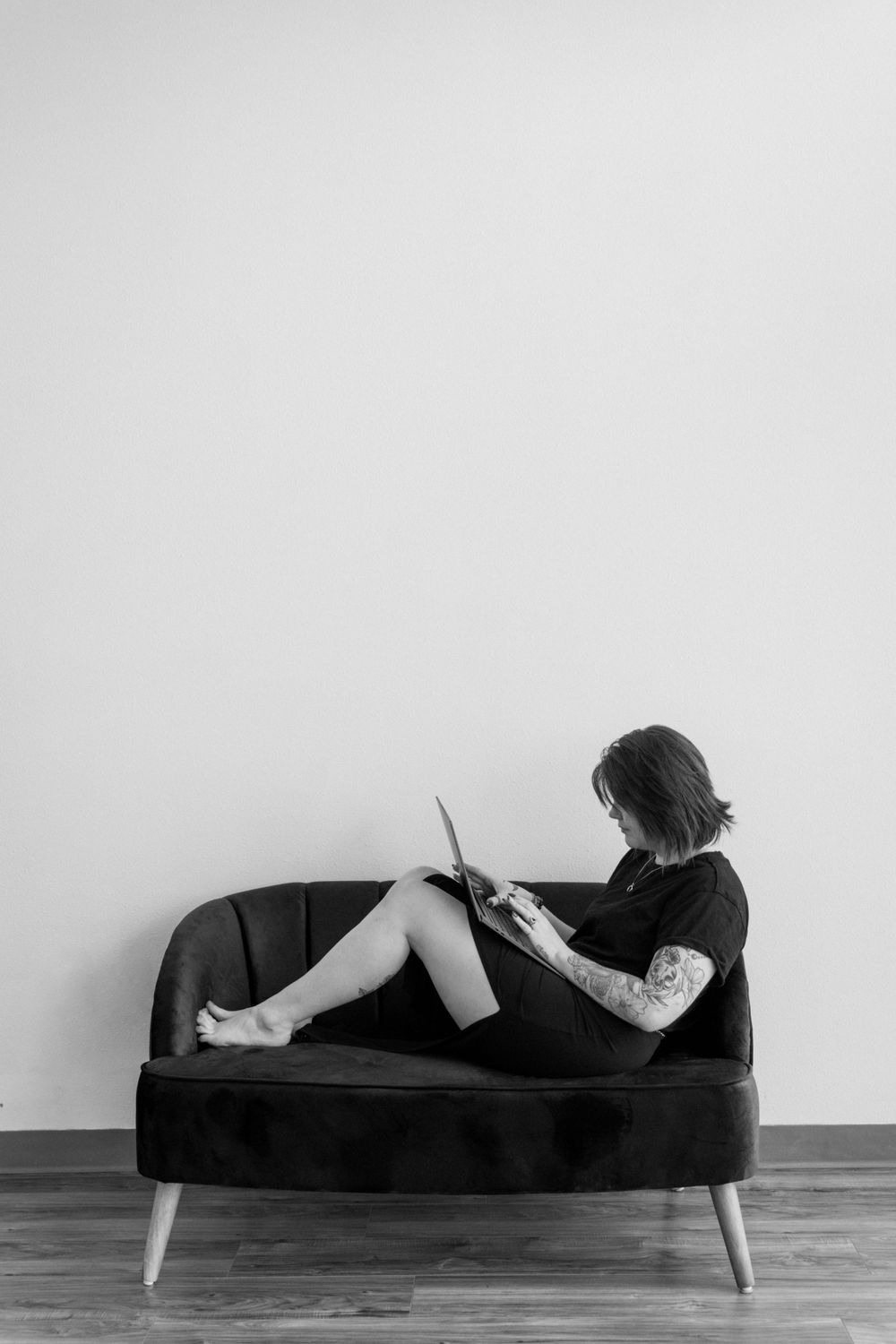 Person in black clothing lounges on a vintage velvet couch while reading in a minimalist black and white setting.