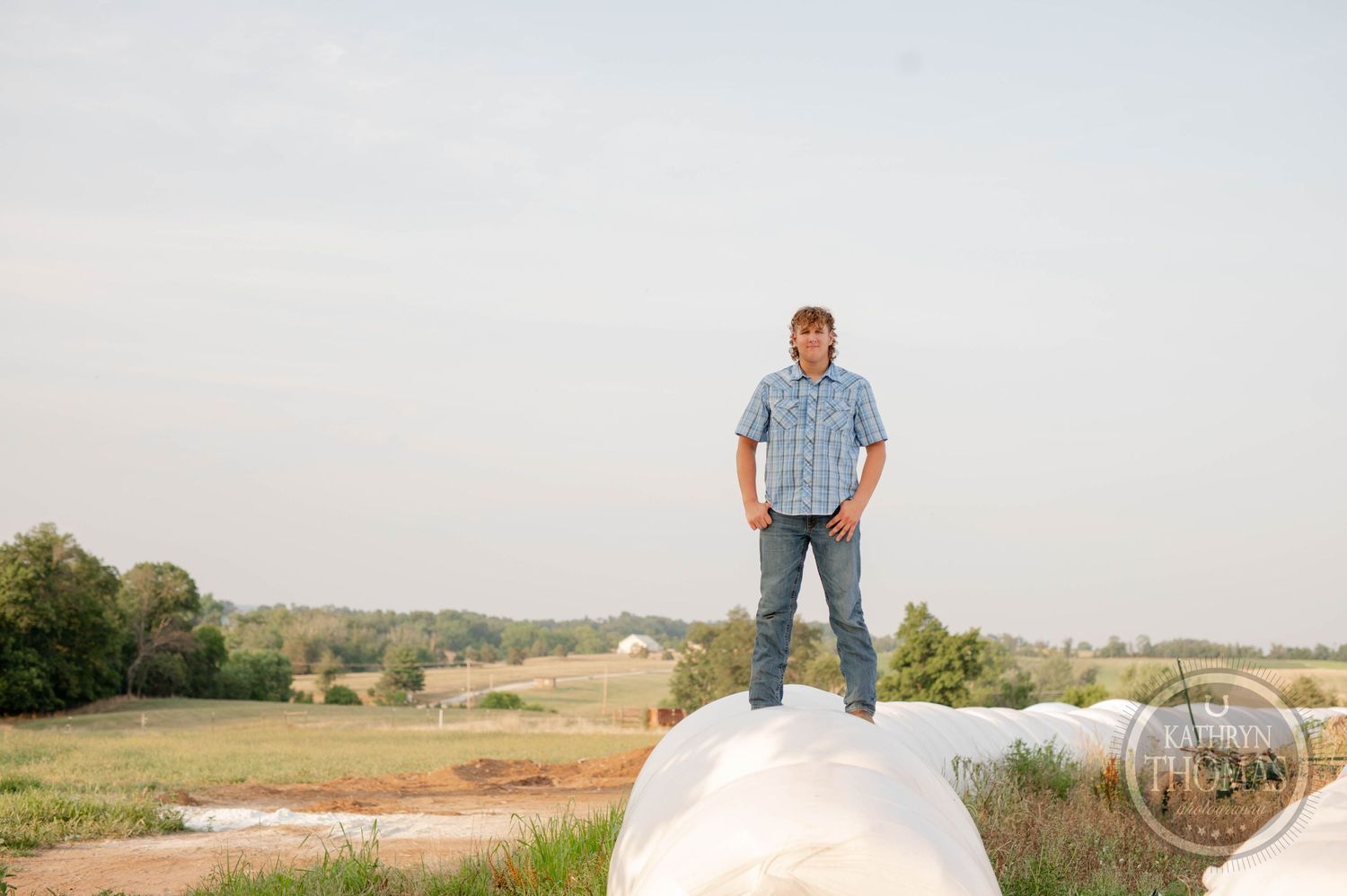 A figure standing confidently atop a large white hay bale in an open field.