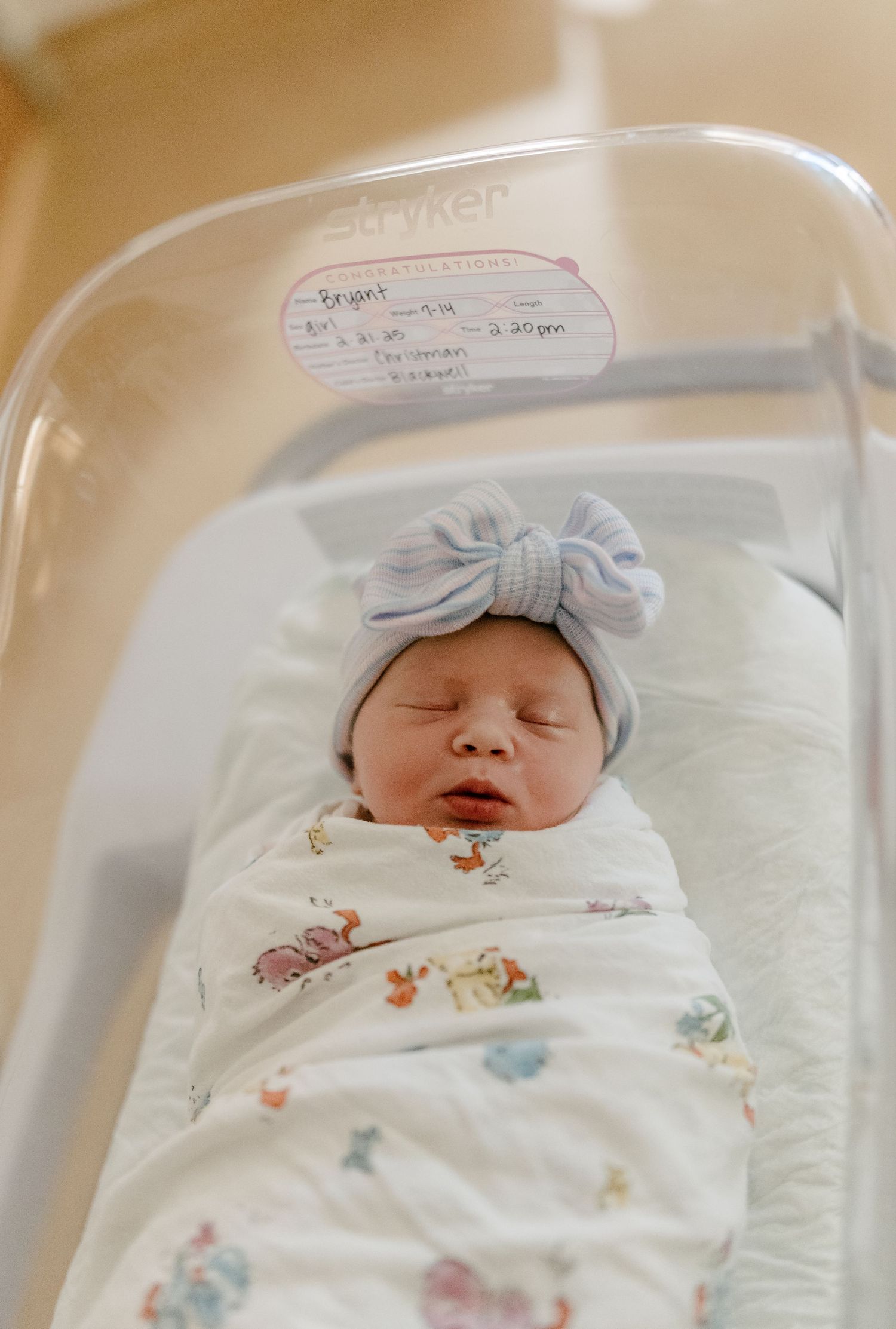 Sleeping newborn wearing soft blue bow hat and white patterned swaddle in hospital bassinet.