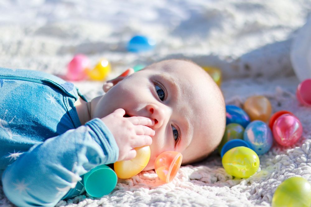 A baby in blue clothing lies on a blanket surrounded by colorful plastic balls and toys.