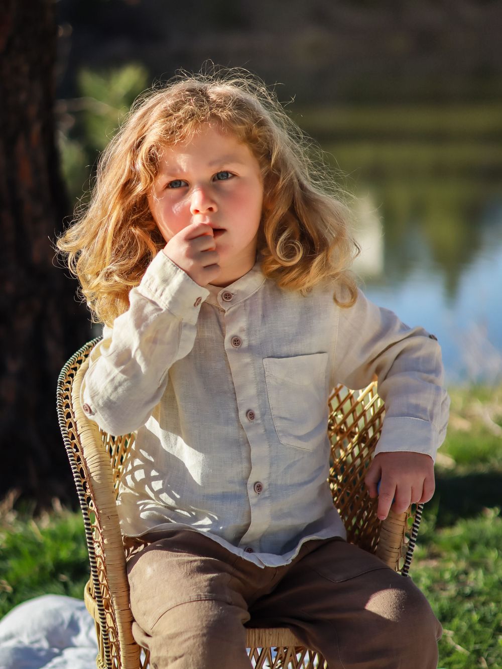 A young child in a white shirt and brown pants sits thoughtfully in an ornate wicker chair by a lake.
