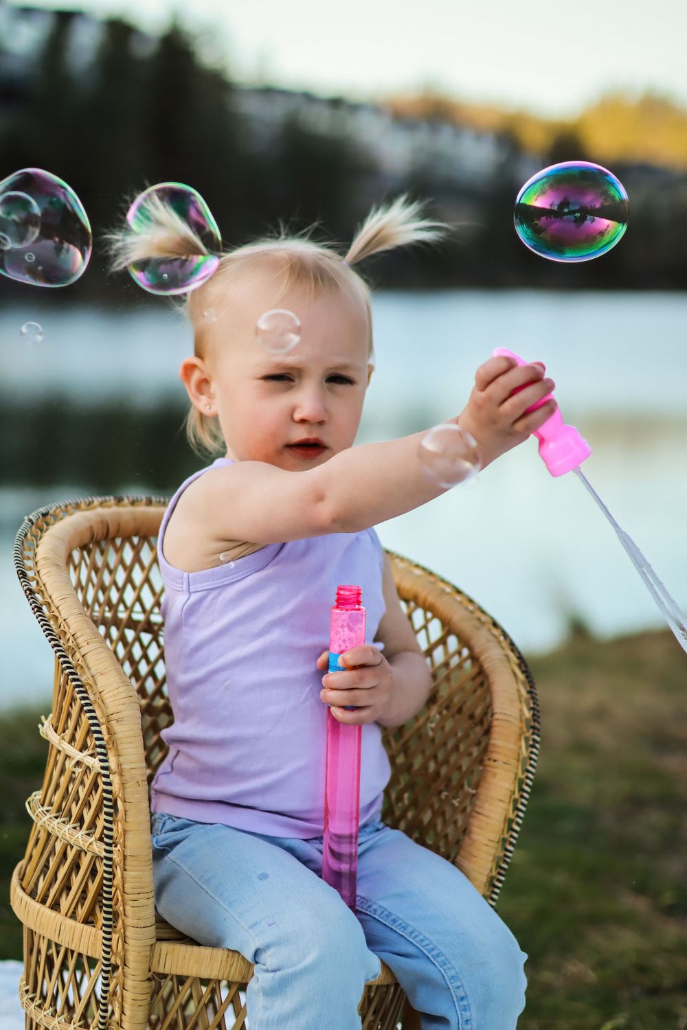 Young child in purple tank top sits in wicker chair blowing bubbles with pink bubble wand by waterfront at sunset.