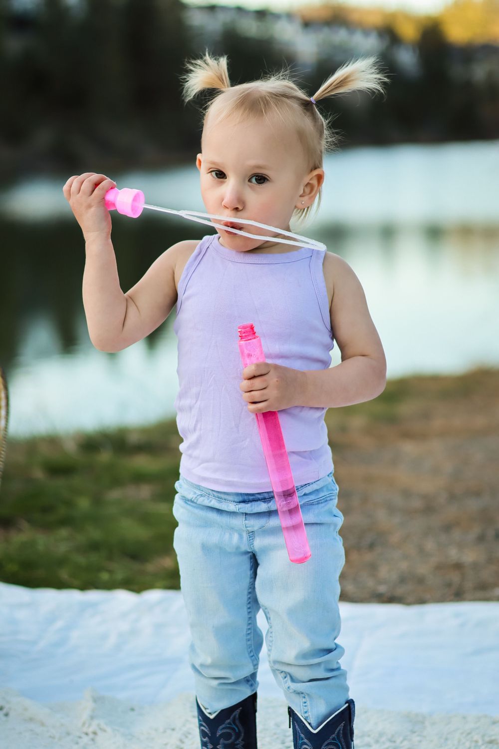 Young child in purple tank top and jeans plays with a pink bubble wand near a lake at sunset.