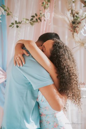 Couple in coordinating blue and floral outfits embrace during a romantic photoshoot with white floral backdrop.