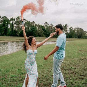 Pink smoke fills the air as couple dances and celebrates in a grassy field by a lake.