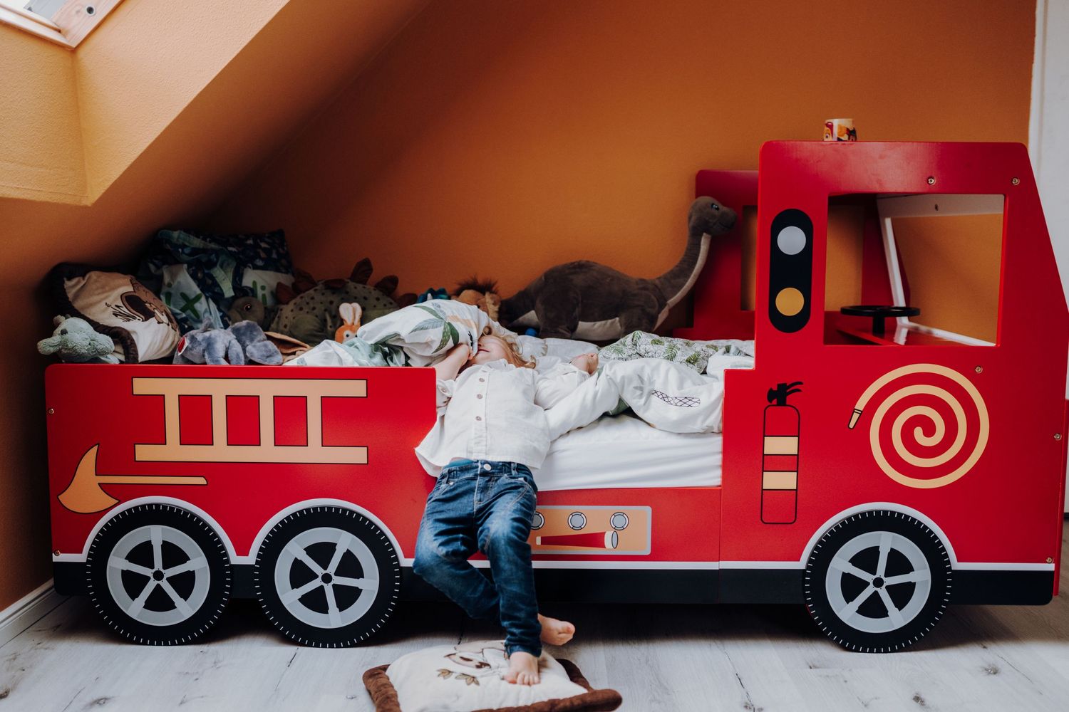 Ein kleiner Junge liegt mit dem Rücken auf seinem Feuerwehrbett und freut sich. Die Wand ist orange und er ist umringt von Kuscheltieren.