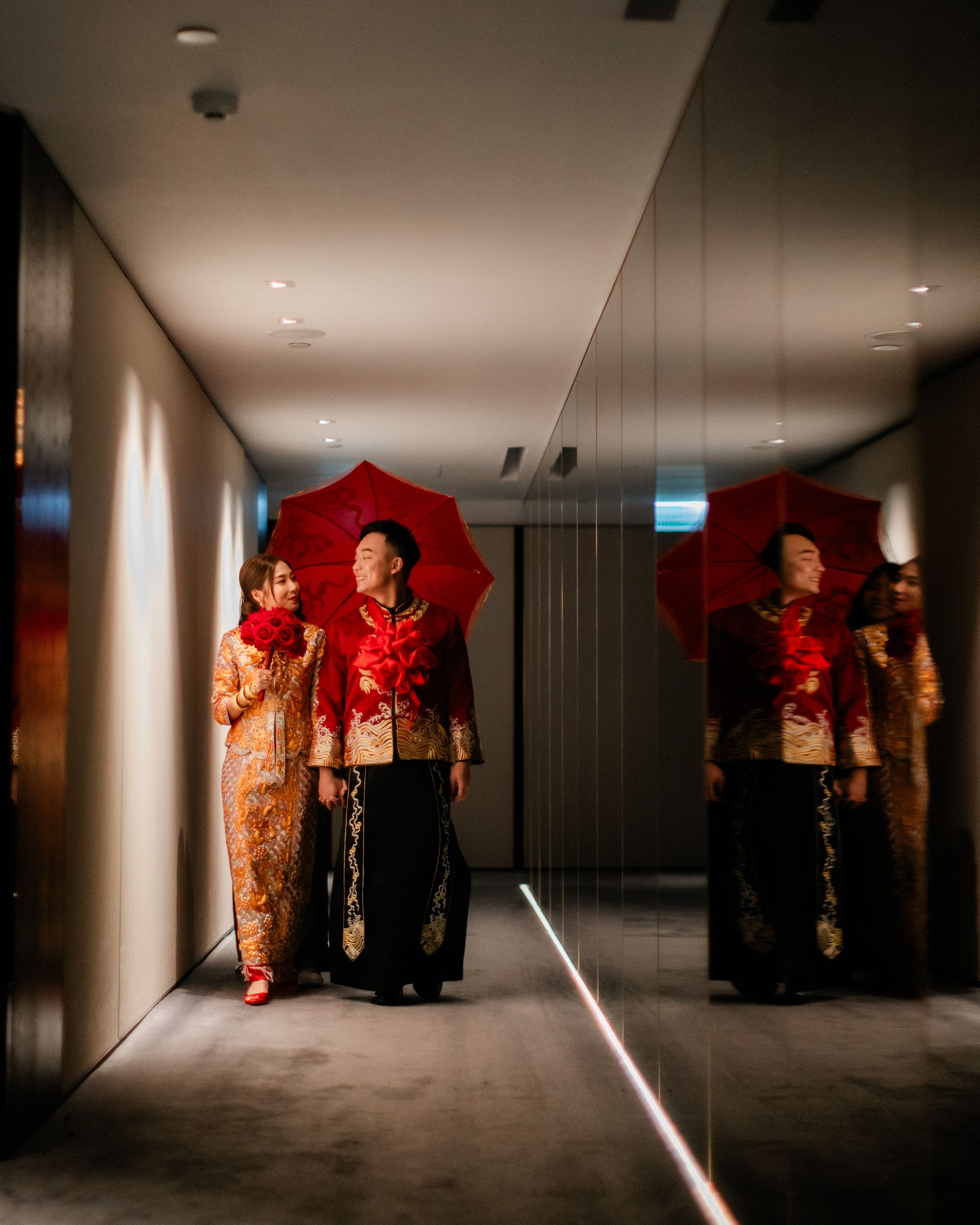Chinese wedding couple in traditional qipao and changshan walk with red umbrella in mirrored hallway.