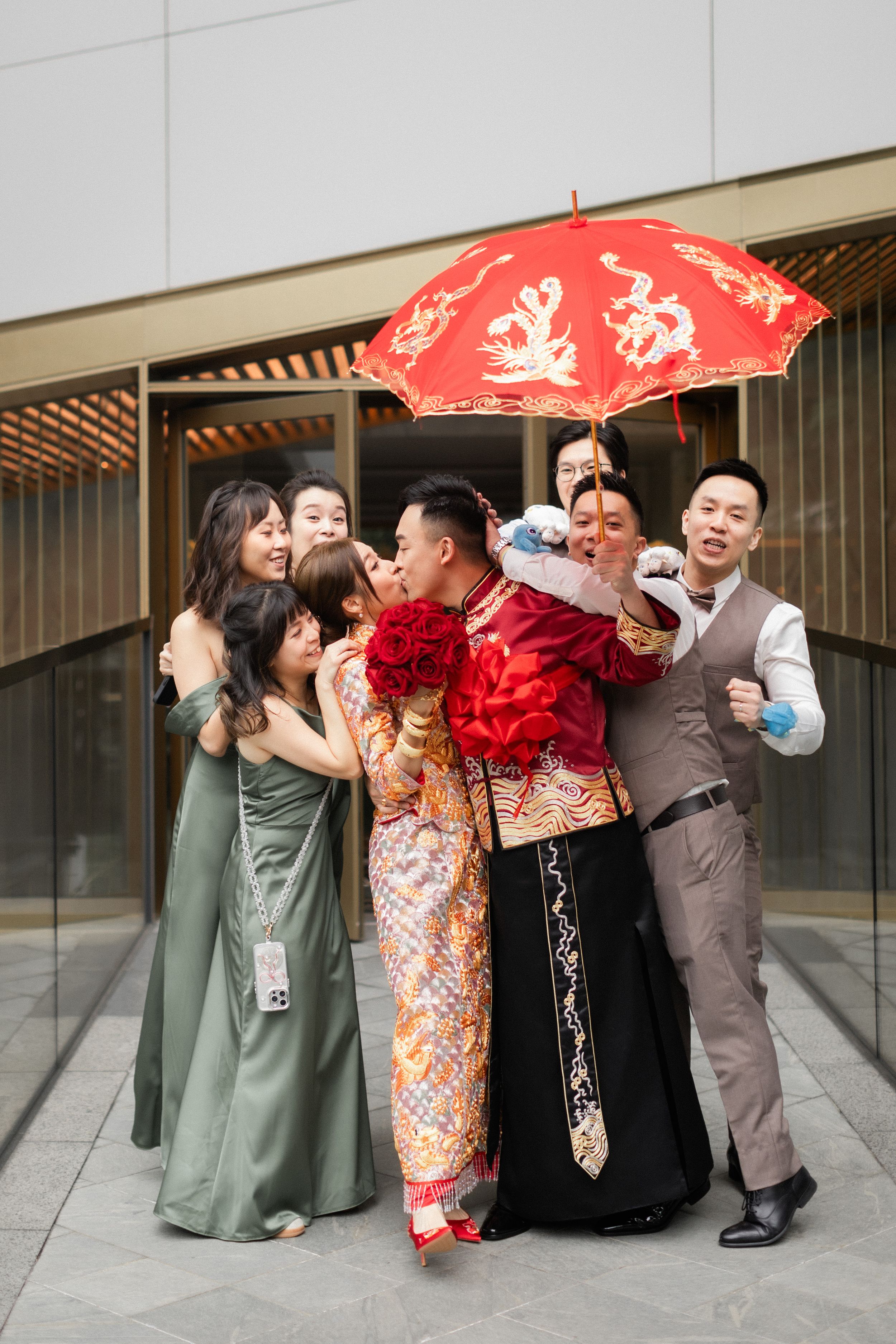 Wedding party hugs Chinese bride and groom under decorative red umbrella outdoors.