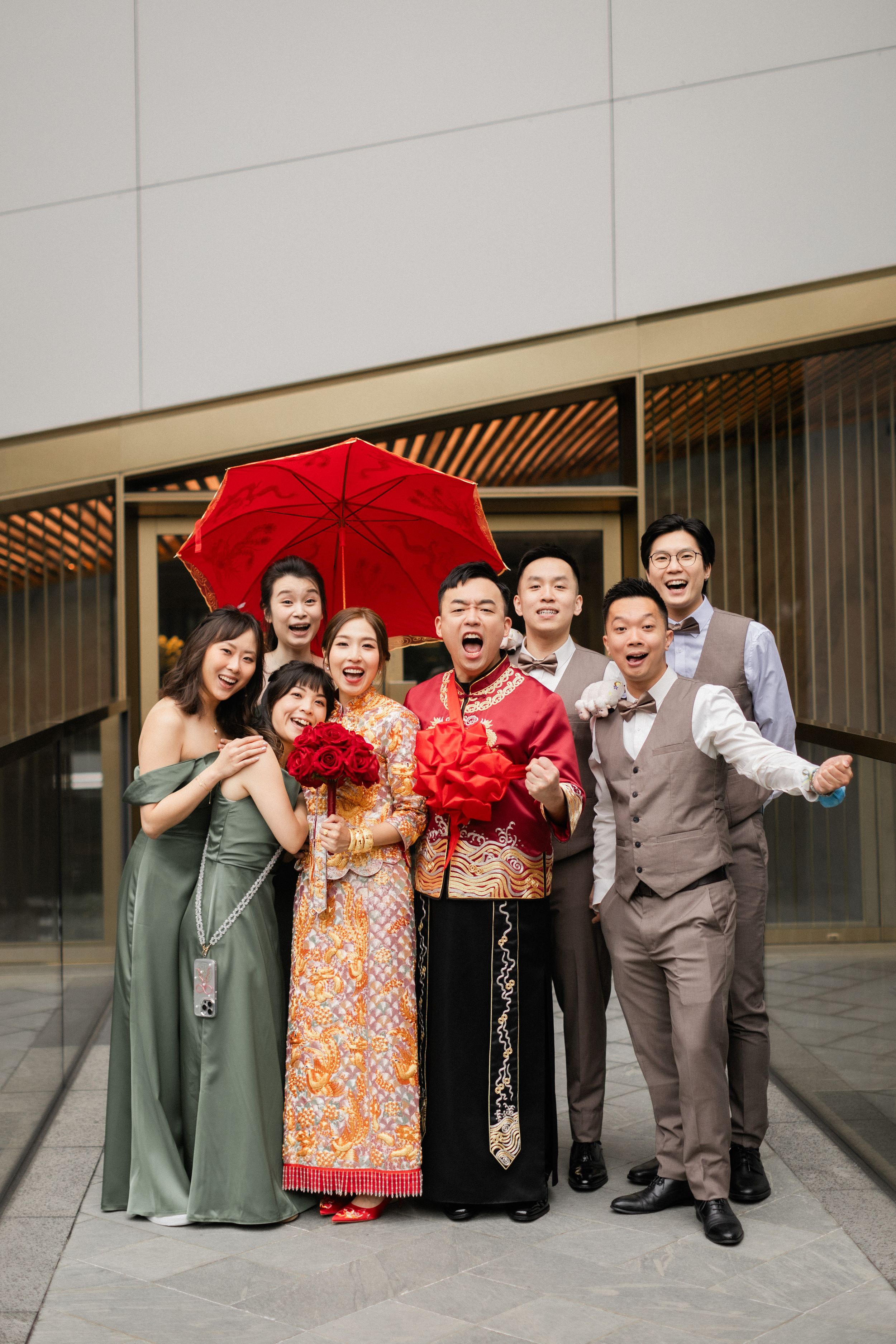 Joyful Chinese wedding party poses under red umbrella in traditional and formal attire.