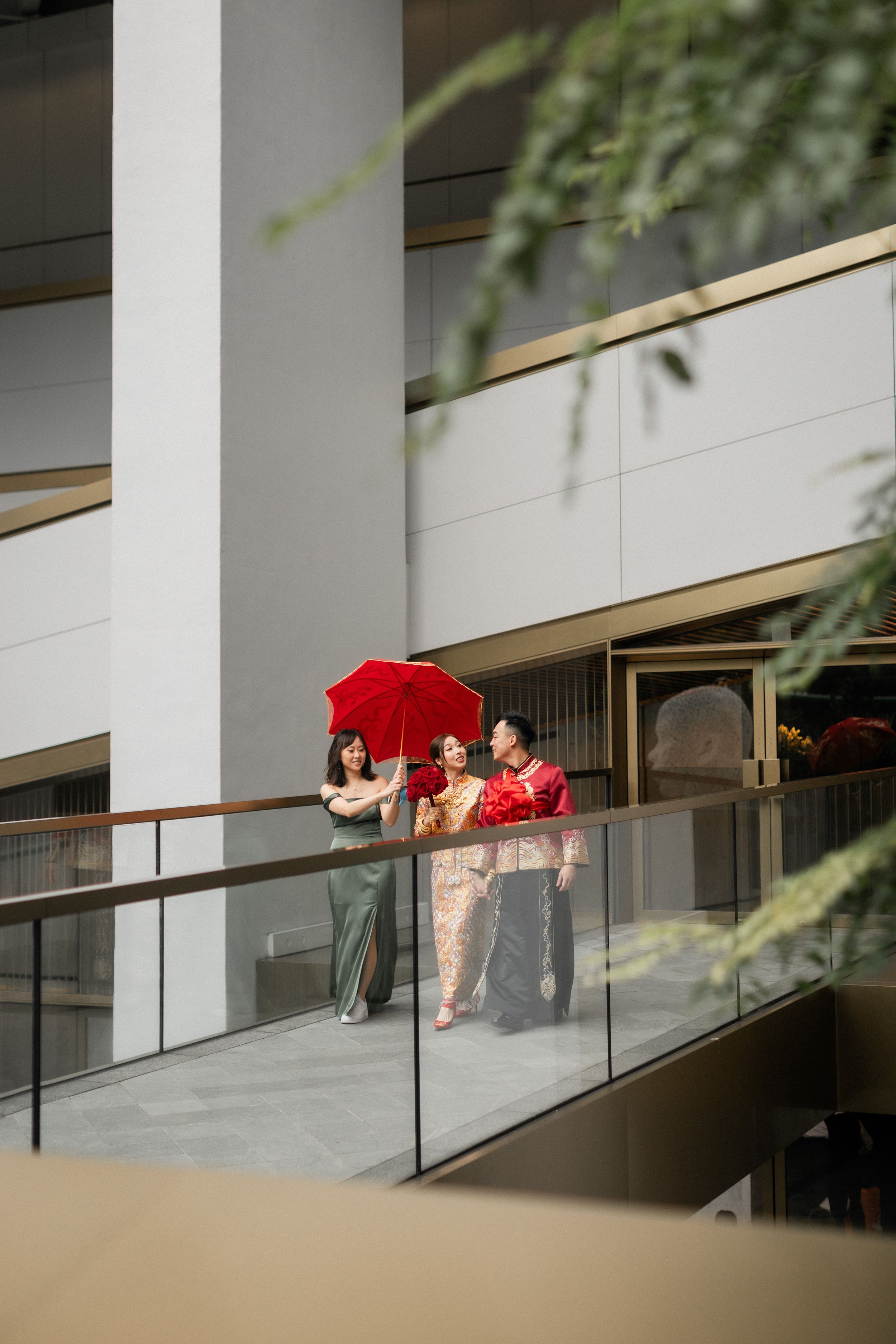 Chinese bride in gold qipao walks with red umbrella on a modern building balcony.
