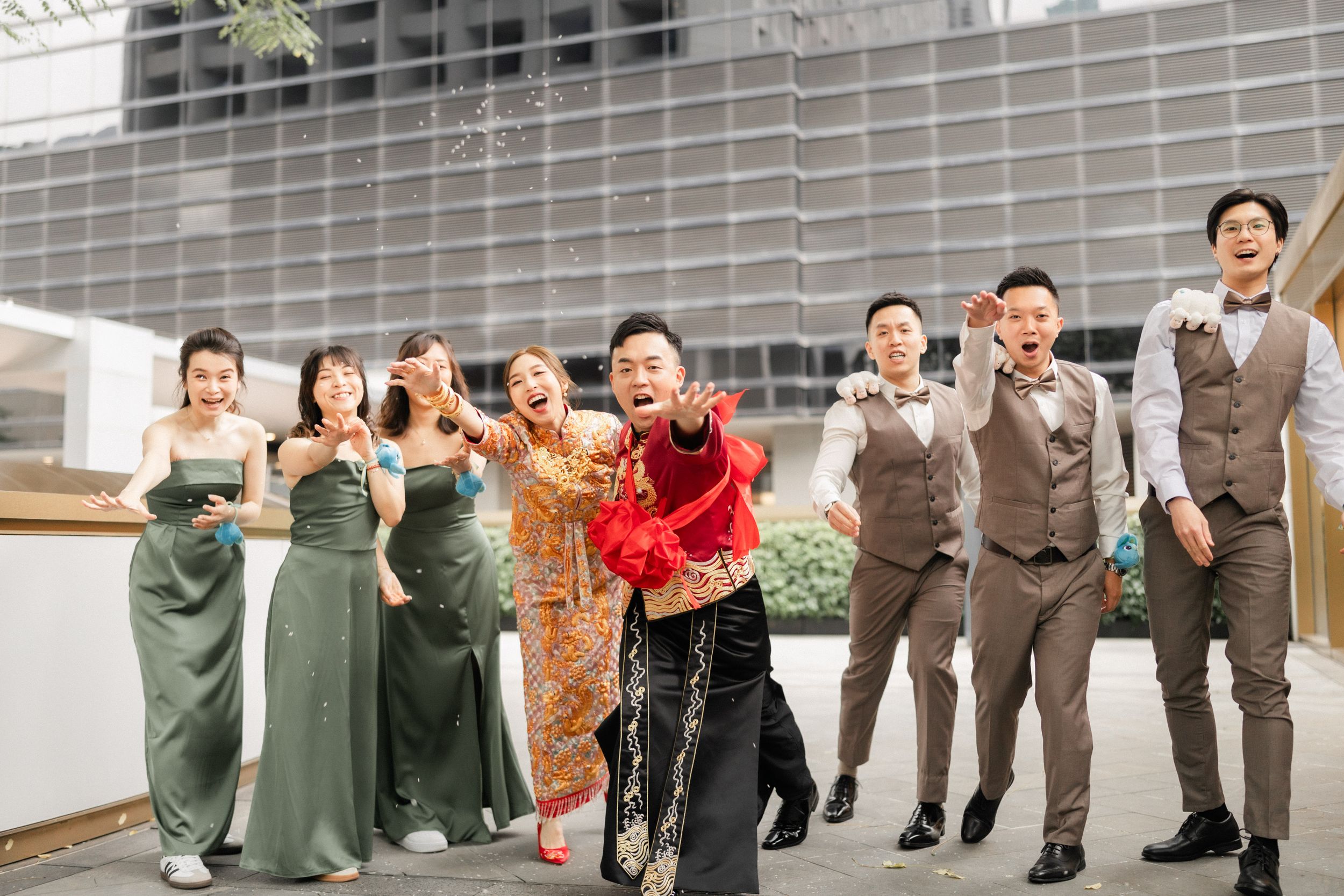 Joyful Chinese wedding party in traditional attire celebrating outdoors with bridesmaids in green dresses and groomsmen in grey suits.