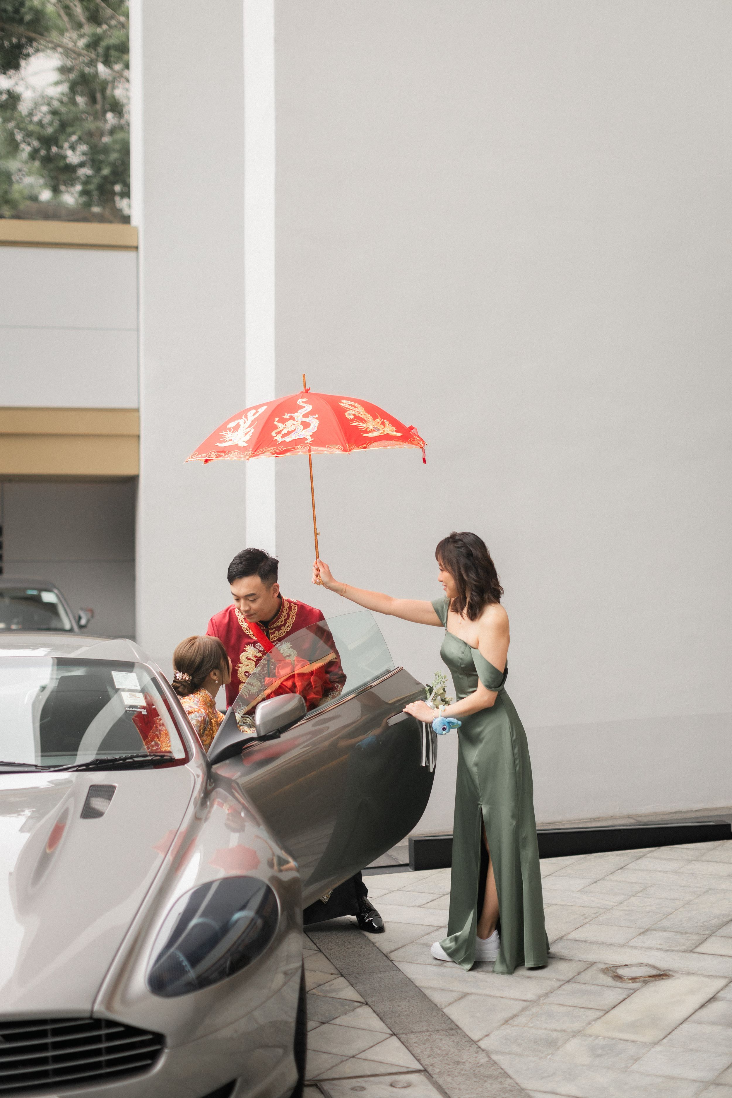 Groom in red Chinese wedding attire exits luxury sports car as bridesmaid holds traditional red double happiness umbrella.