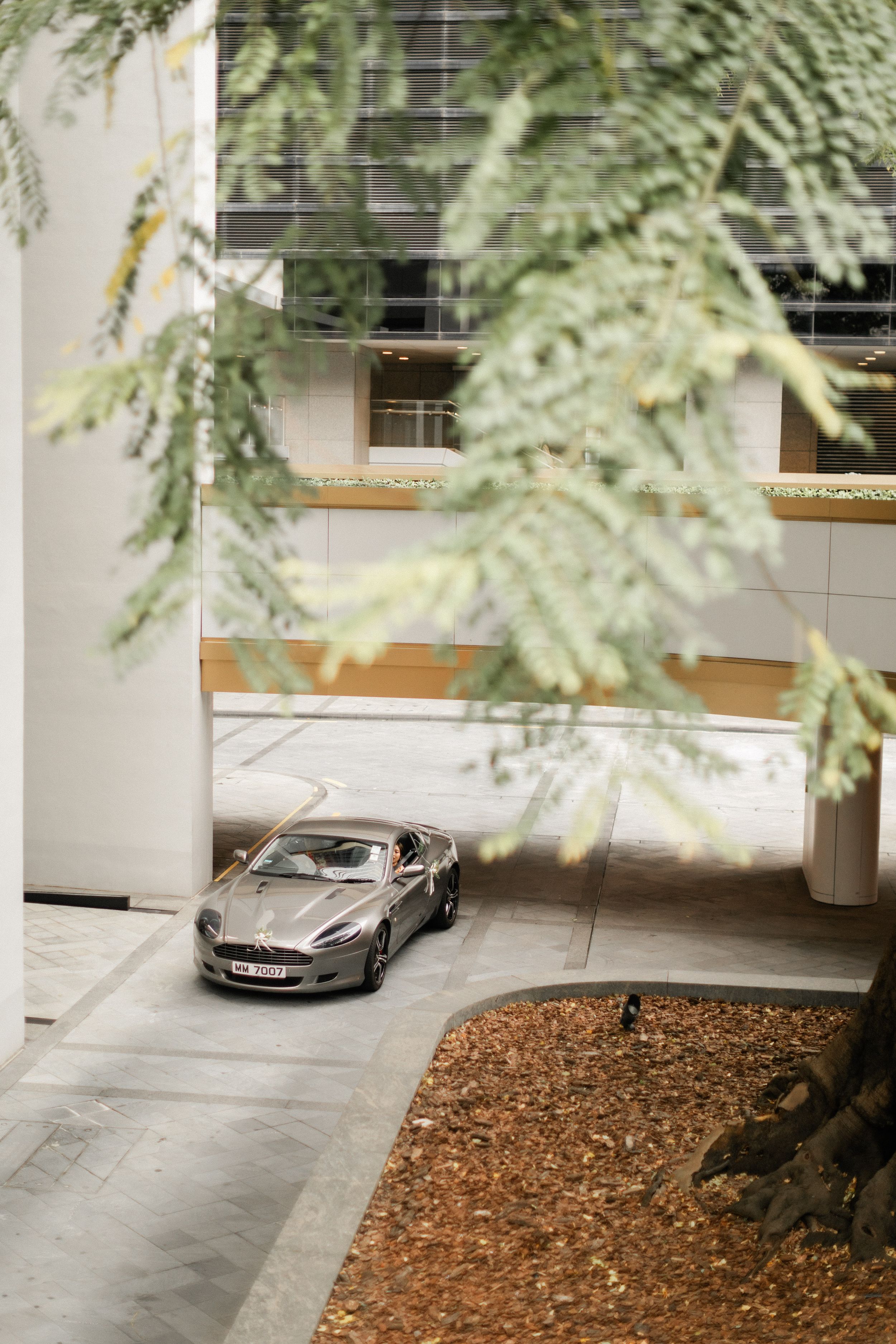 Aerial view of silver Aston Martin luxury sports car parked in modern building courtyard framed by green tree branches.