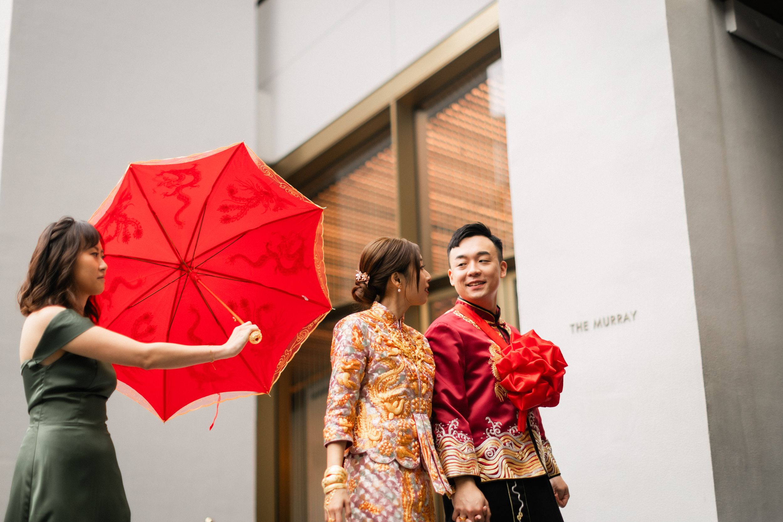 Chinese wedding couple in traditional red and gold qipao attire outside The Murray hotel with red ceremonial umbrella.